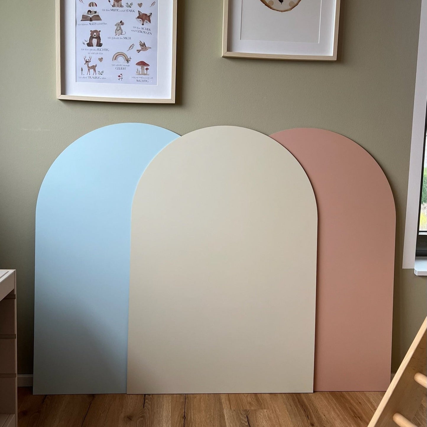 Three arch-shaped decorative boards in soft pastel colors: blue, beige, and pink, displayed on a wall.